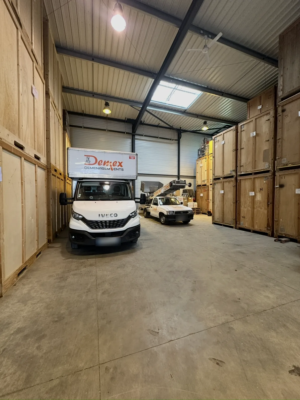 Entrepôt de garde-meubles DEMEX : conteneurs en bois empilés de chaque côté, camion DEMEX Déménagements au centre