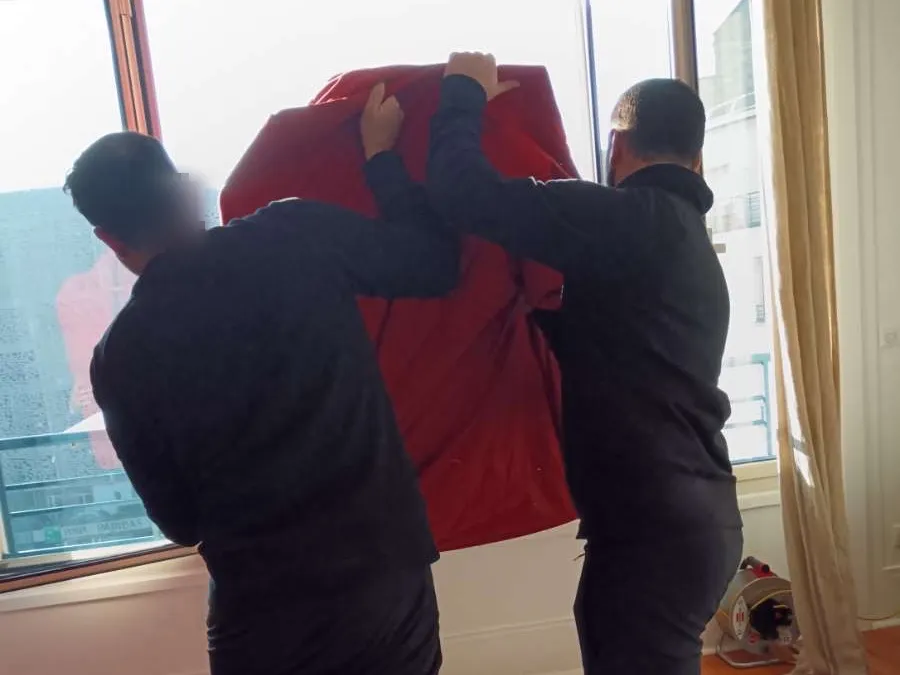Deux déménageurs DEMEX soulevant un objet volumineux emballé dans une housse de protection rouge
